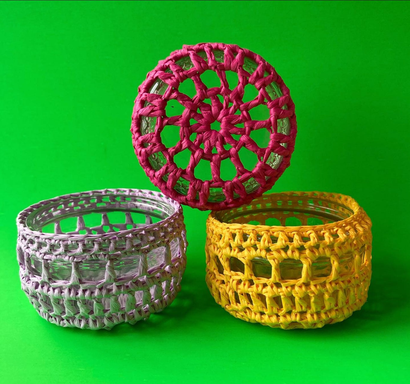 Alt text: the photo shows 3 upcycled tealight holders made using raffia crocheted over Gu pots. The left pot is lilac, the right is bright yellow, and the top one is hot pink, resting on the side showing the design on the bottom of the pot. 